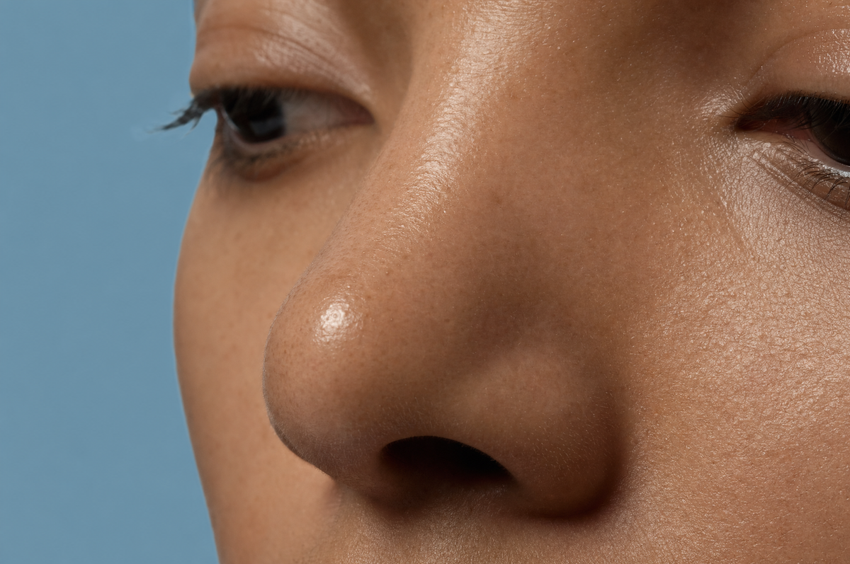 Editorial dusty-blue close-up of an adult's nose and the center of the face, where glasses rest and breakouts cluster