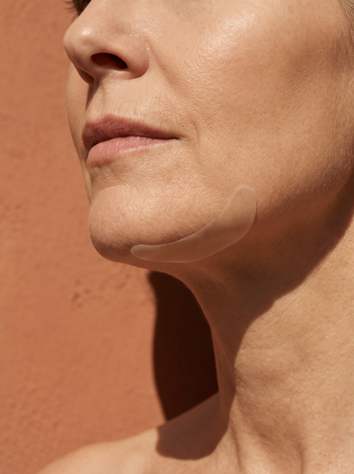 Close-up of an adult chin and jawline wearing a crescent-shaped, nearly invisible hydrocolloid patch on a terracotta background