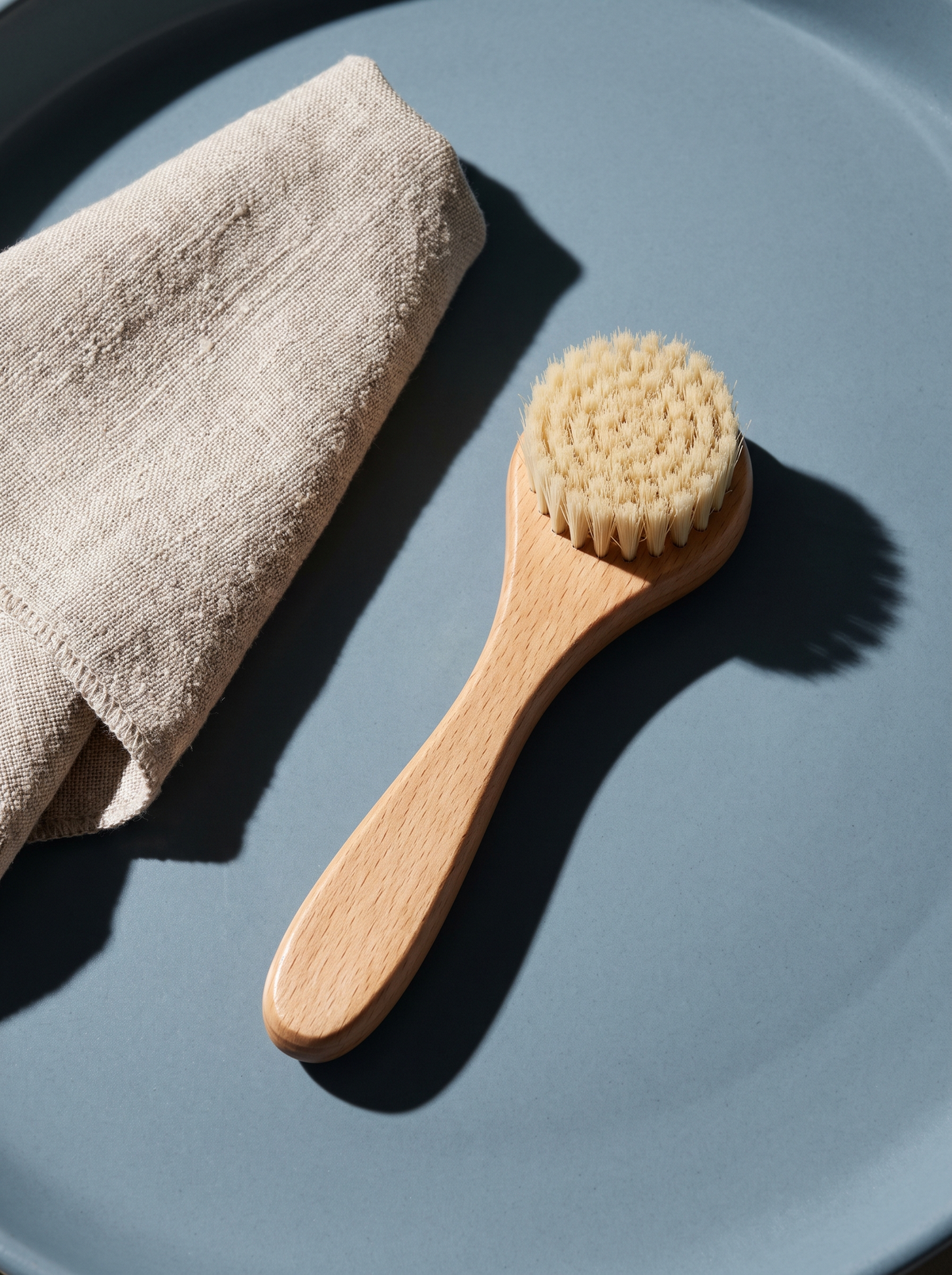 A facial cleansing brush beside a textured washcloth on a dusty-blue surface