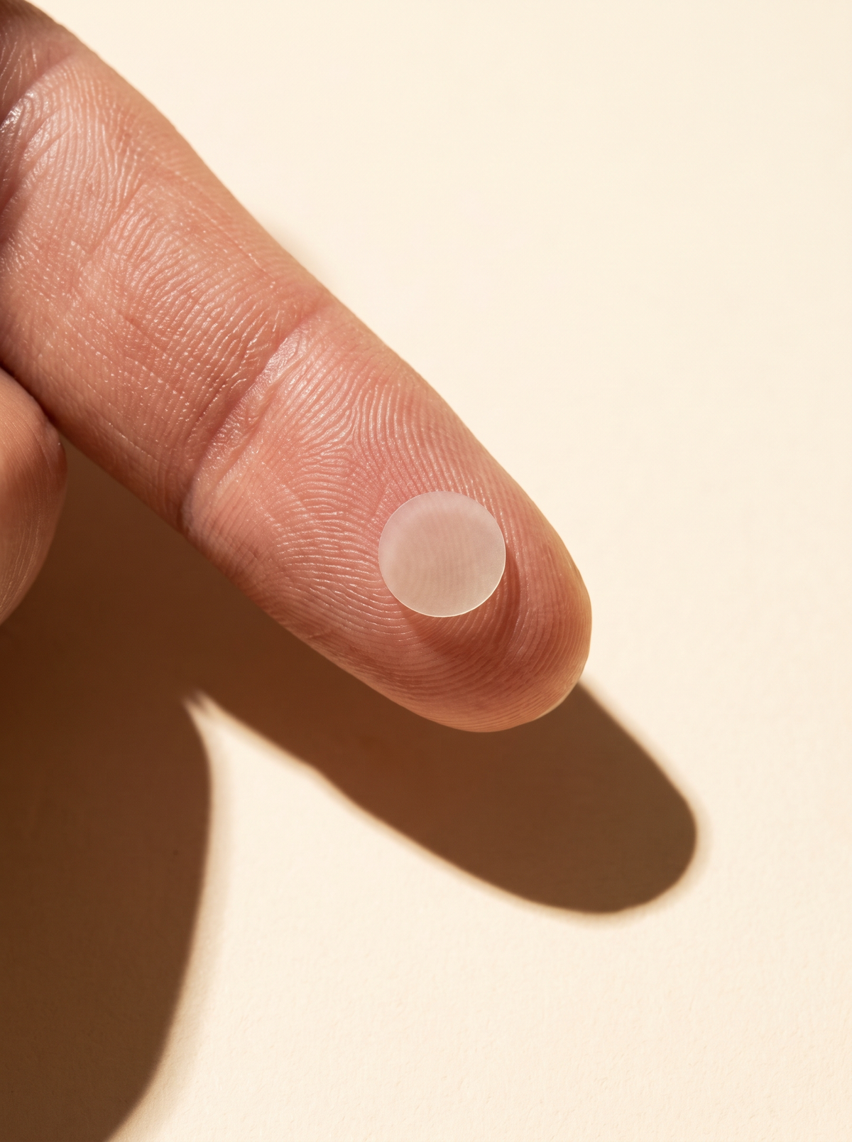 A single clear hydrocolloid dot held on an adult's fingertip against warm cream