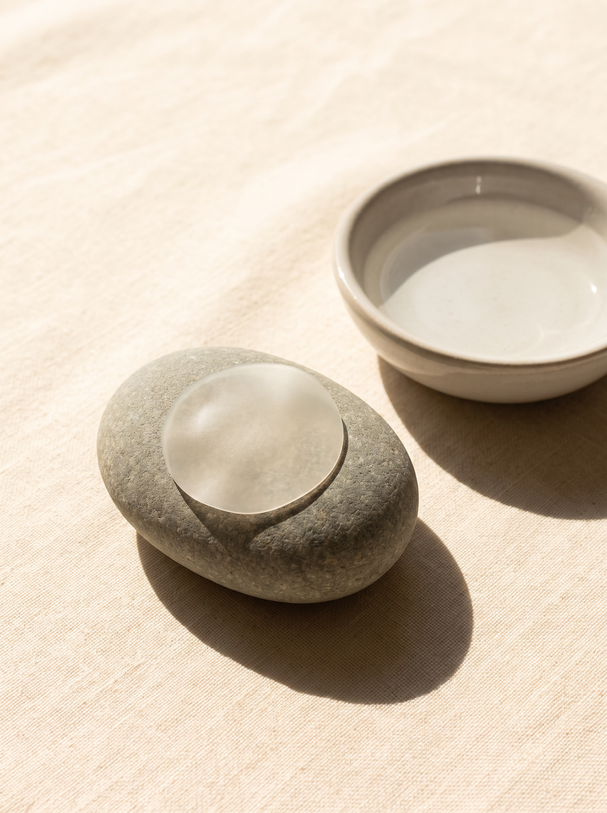 A single thin, nearly invisible hydrocolloid dot on a smooth grey river stone beside a shallow dish of water on warm cream