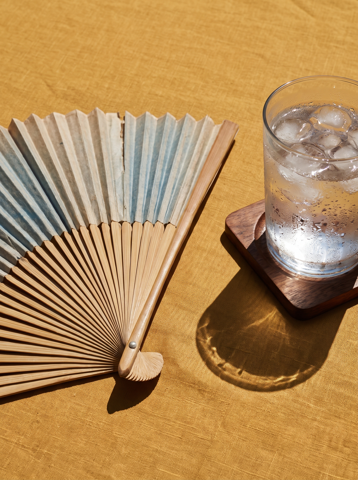 A folding hand fan beside a glass of ice water on a golden ochre surface in strong sun