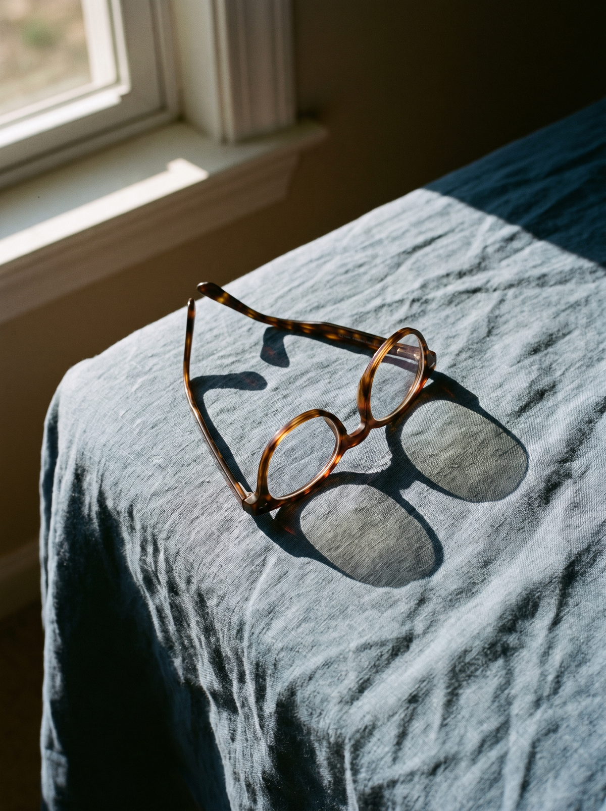 Tortoiseshell glasses folded on dusty-blue linen, resting where nose pads press the sides of the nose
