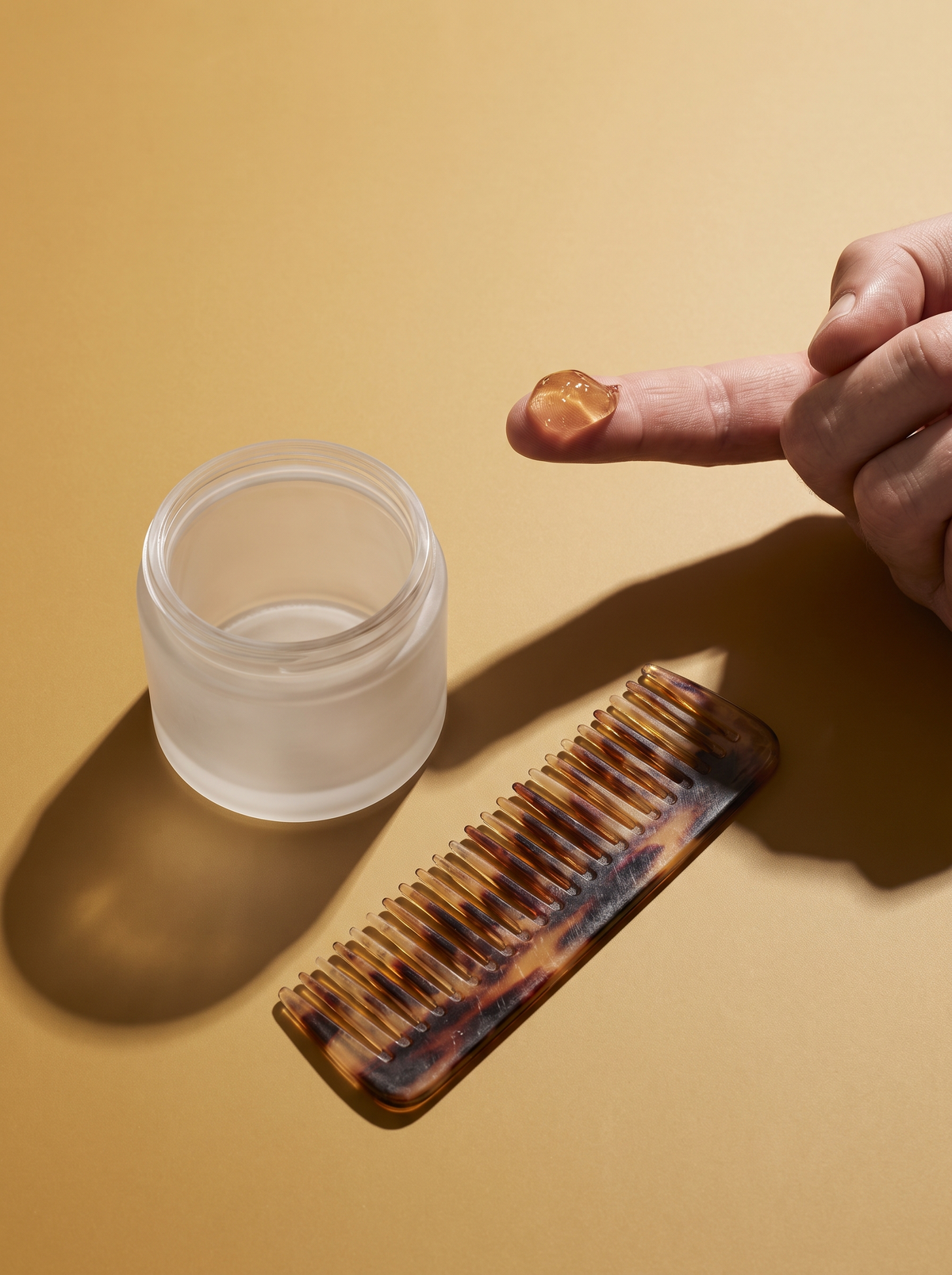 A plain tub of hair styling gel with a dollop on an adult fingertip beside a wide-tooth comb on a golden ochre surface