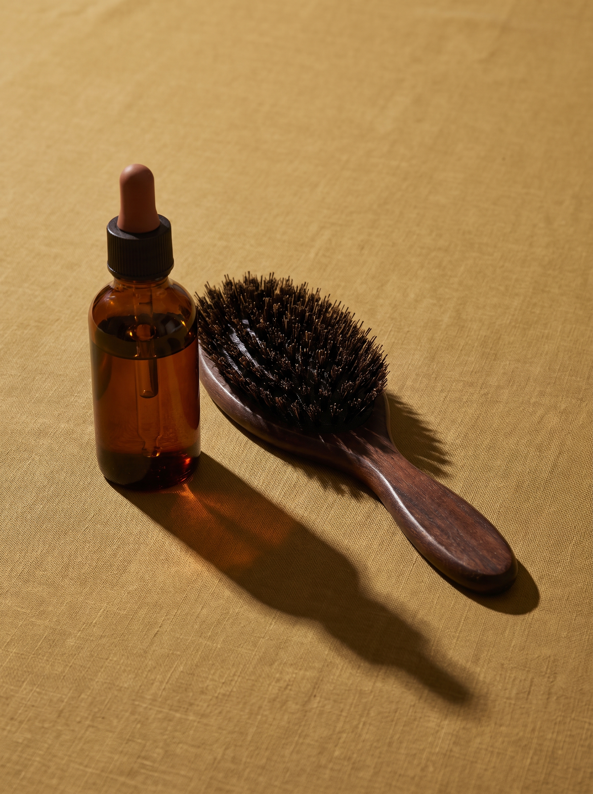 A small amber bottle of hair oil with a dropper and a wooden hairbrush on a golden ochre surface
