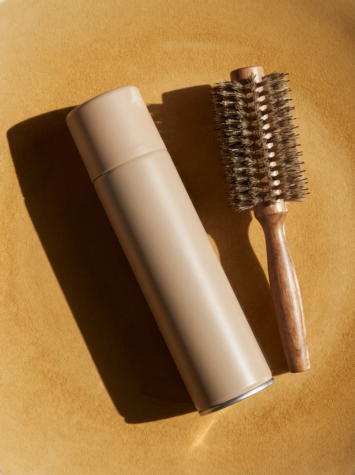 A plain hair spray can and a round styling brush on a golden ochre surface