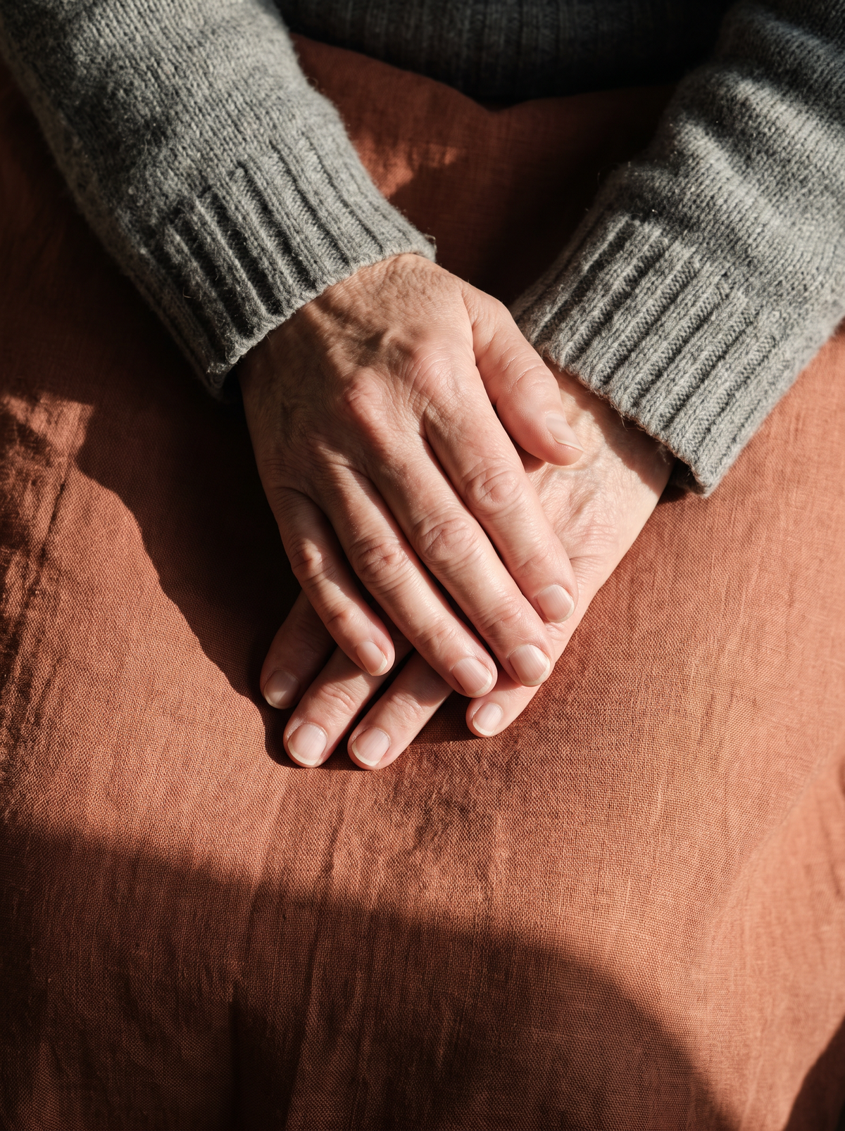 An adult's hands resting calmly folded in their lap with soft knit sleeves on terracotta linen