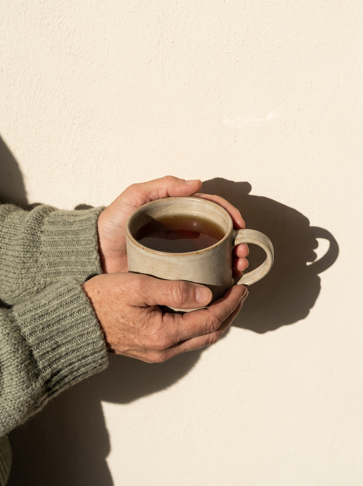 An adult's hands in sage-green knit sleeves wrapped around a mug of tea