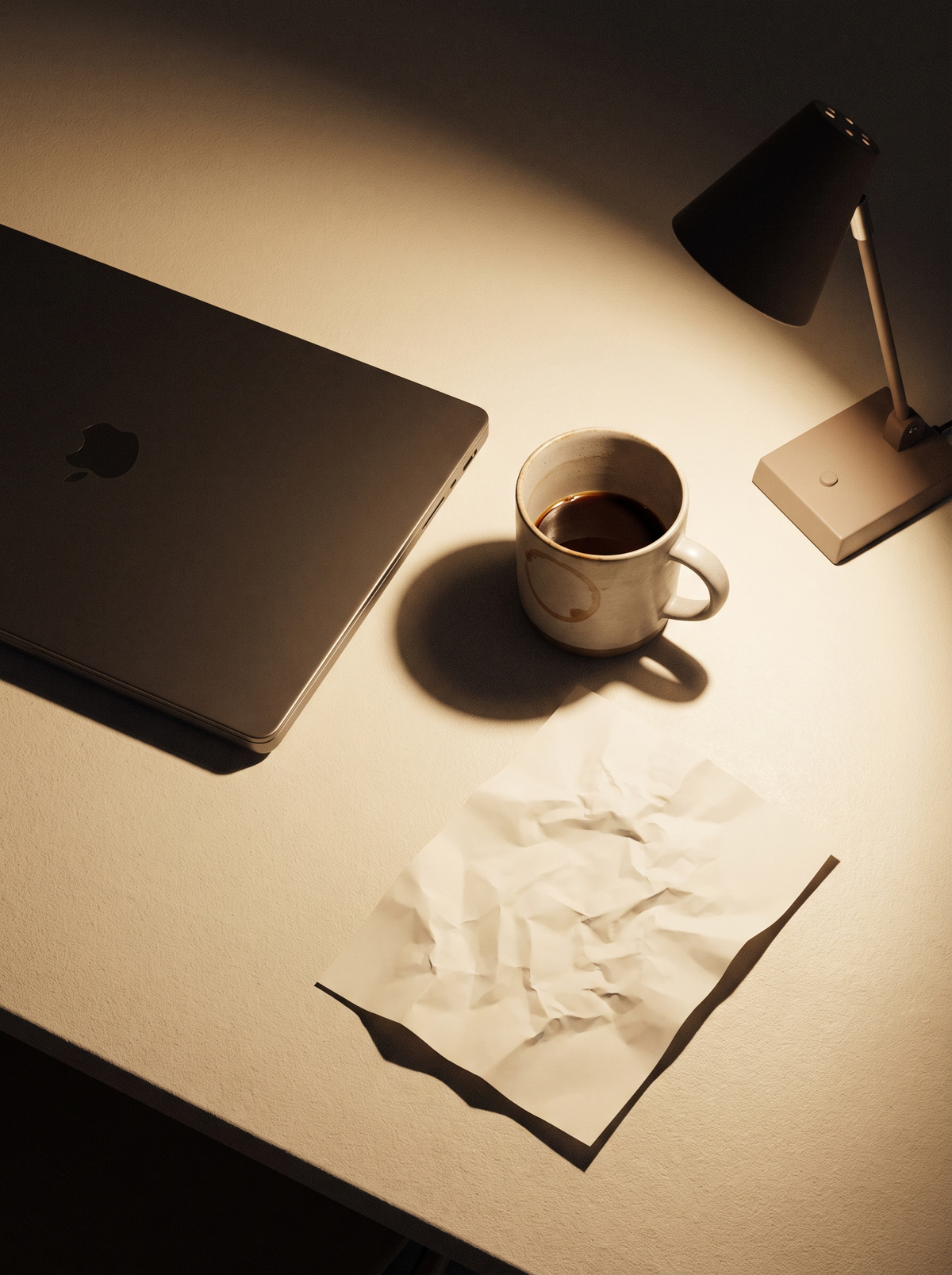 A late-night desk with a closed laptop, a mug of coffee, crumpled paper and a small lamp