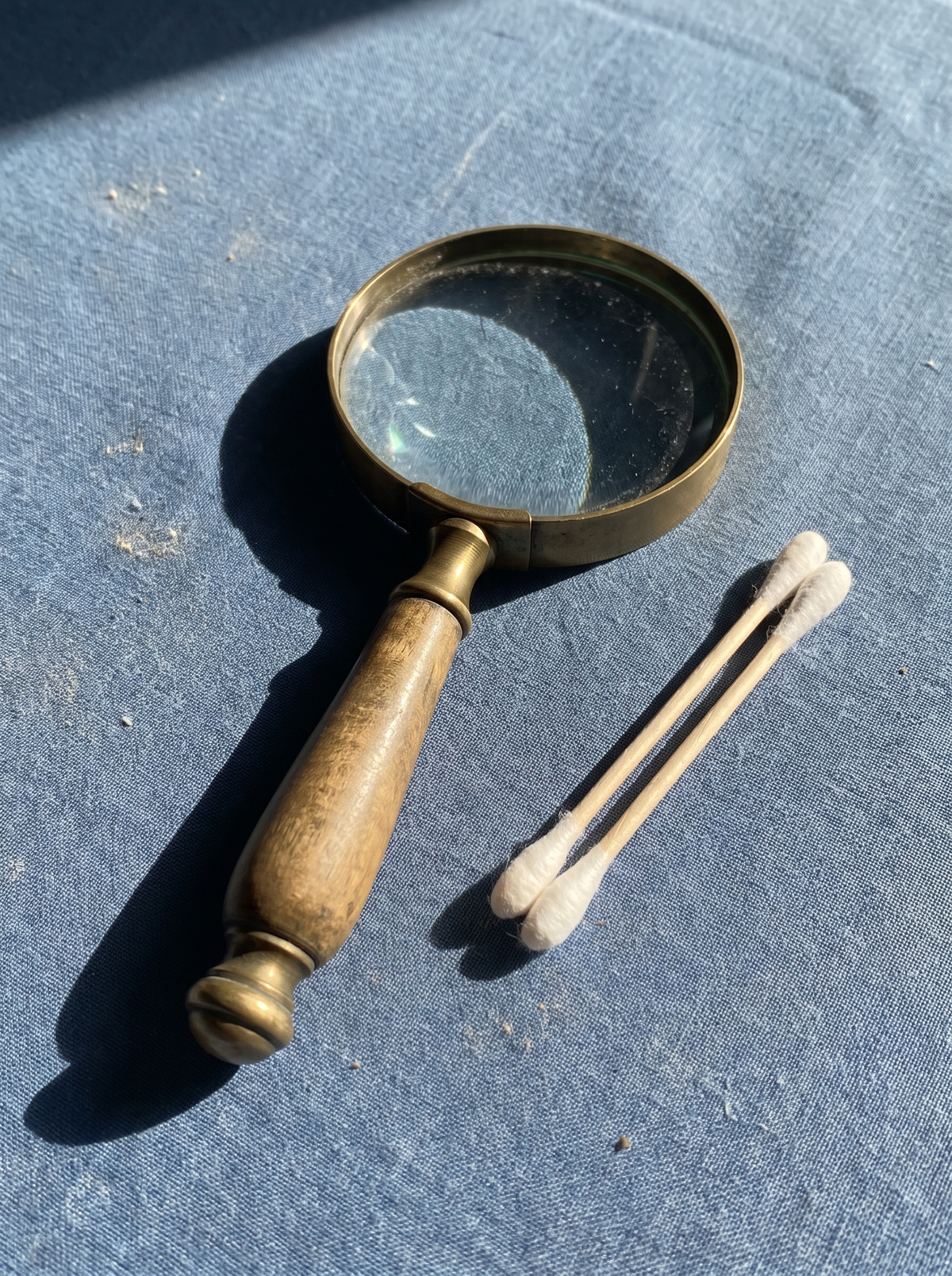 A brass-handled magnifying glass beside two cotton swabs on a dusty-blue surface